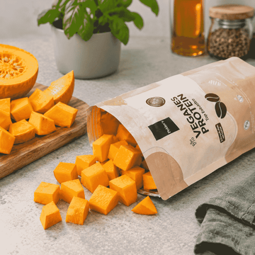 Im Hintergrund ist ein aufgeschnittener Kürbis zu erkennen. Im Vordergrund fallen gewürfelte Kürbisstücke aus einer Pelsters-Protein-Verpackung.